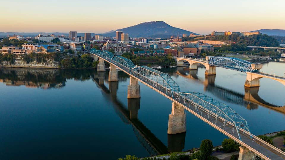 A steel bridge spanning a calm river, with a cityscape and mountains in the background.
