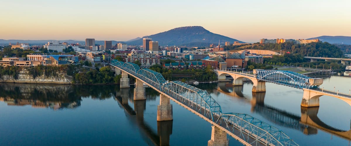 A steel bridge spanning a calm river, with a cityscape and mountains in the background.