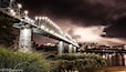 Un puente iluminado con base de piedra, un río y un paisaje urbano al fondo.