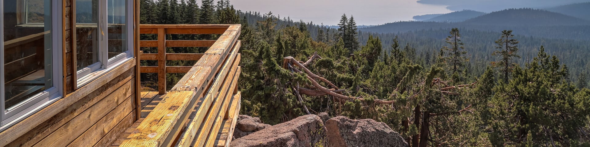 A wooden cabin with a wooden deck overlooking a forested valley and a lake.