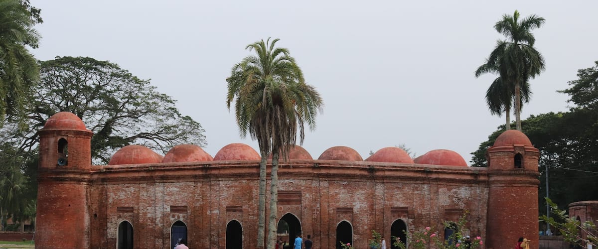 A historic building with red domes and arched doorways, surrounded by palm trees and greenery.