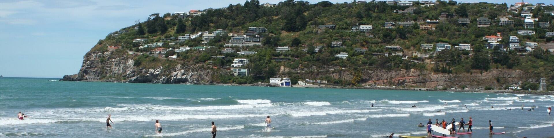 A beach with waves, rocks, and houses on a hillside in the background.