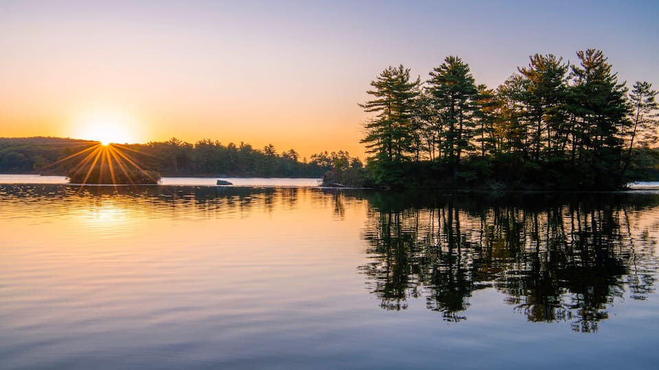 A calm lake at sunset with reflections of trees and a clear sky.