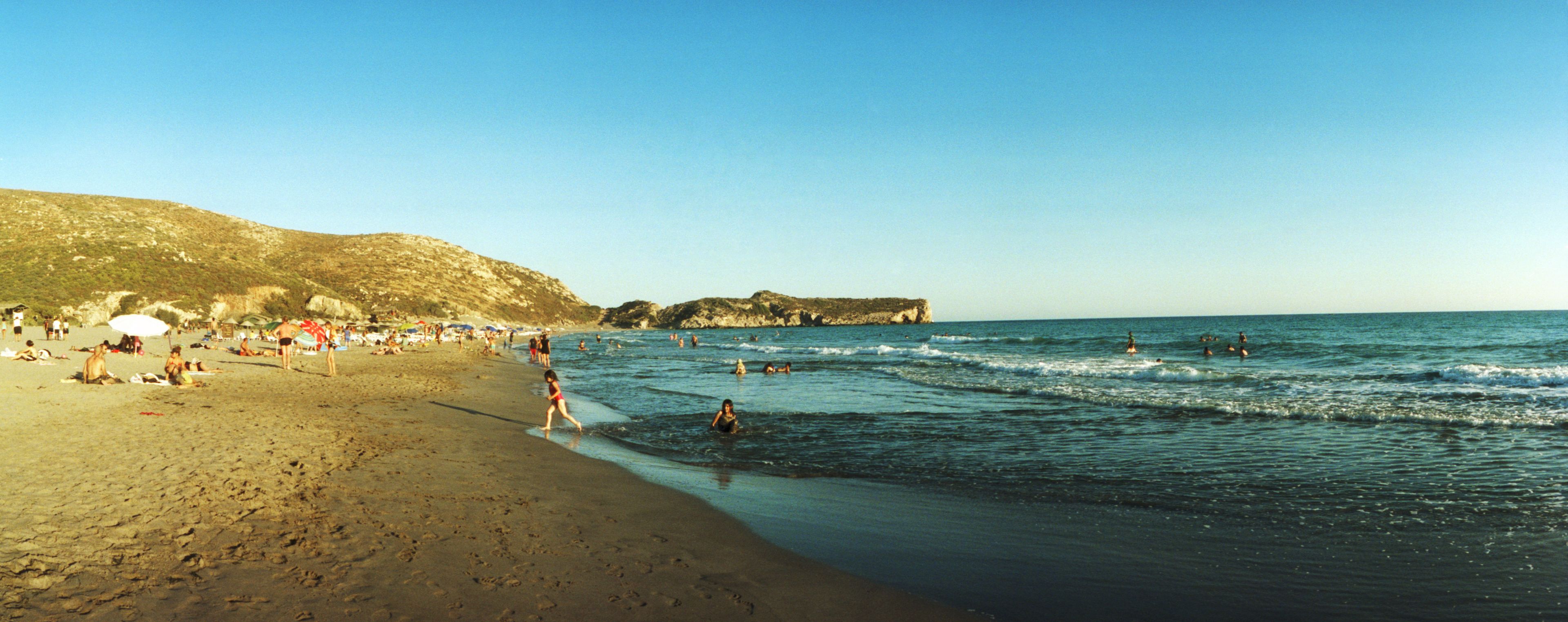 Une plage où l’on voit des gens se baigner et se promener le long du rivage, une côte rocheuse et des parasols plantés dans le sable.