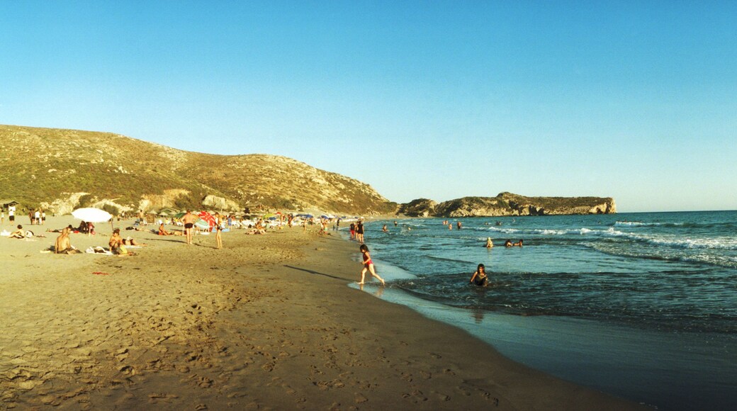 Une plage où l’on voit des gens se baigner et se promener le long du rivage, une côte rocheuse et des parasols plantés dans le sable.