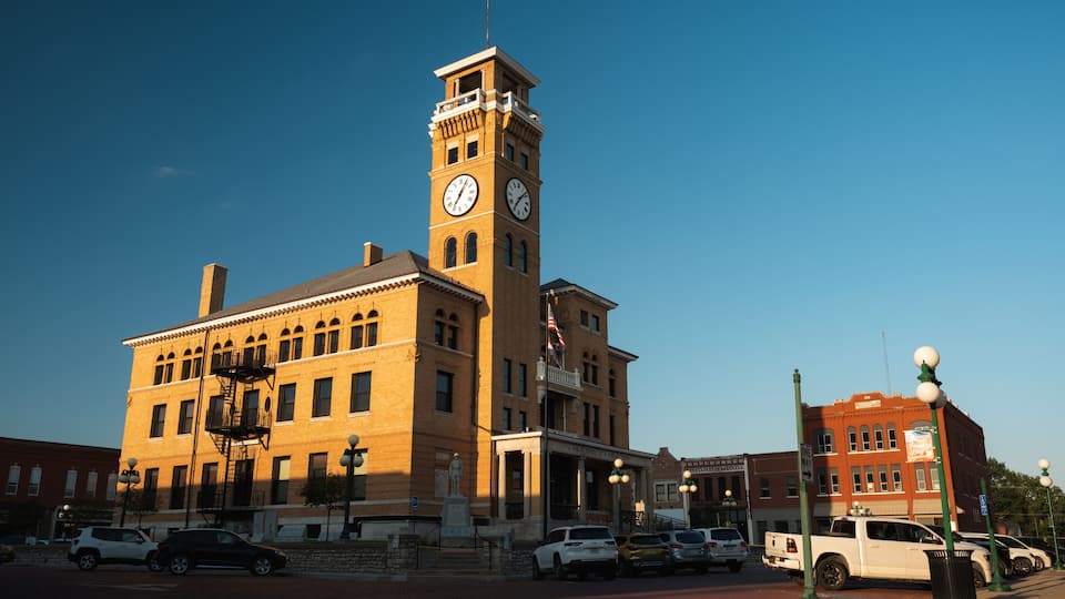A historic building with a clock tower, a statue, and a red brick plaza with parked cars.