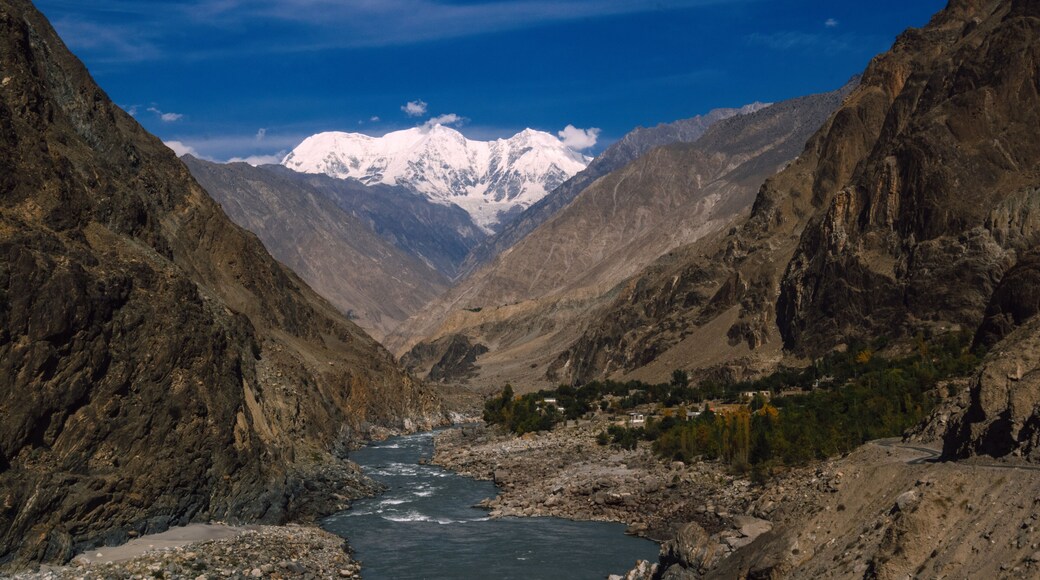 View of a glacial river cutting through a rugged valley, framed by towering, rocky mountains under a vibrant blue sky, with a snow-capped peak in the distance, Jaglot, Gilgit Baltistan, Pakistan.