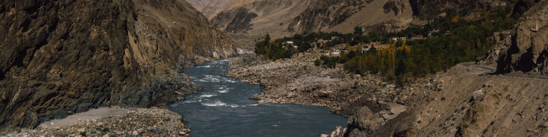 View of a glacial river cutting through a rugged valley, framed by towering, rocky mountains under a vibrant blue sky, with a snow-capped peak in the distance, Jaglot, Gilgit Baltistan, Pakistan.