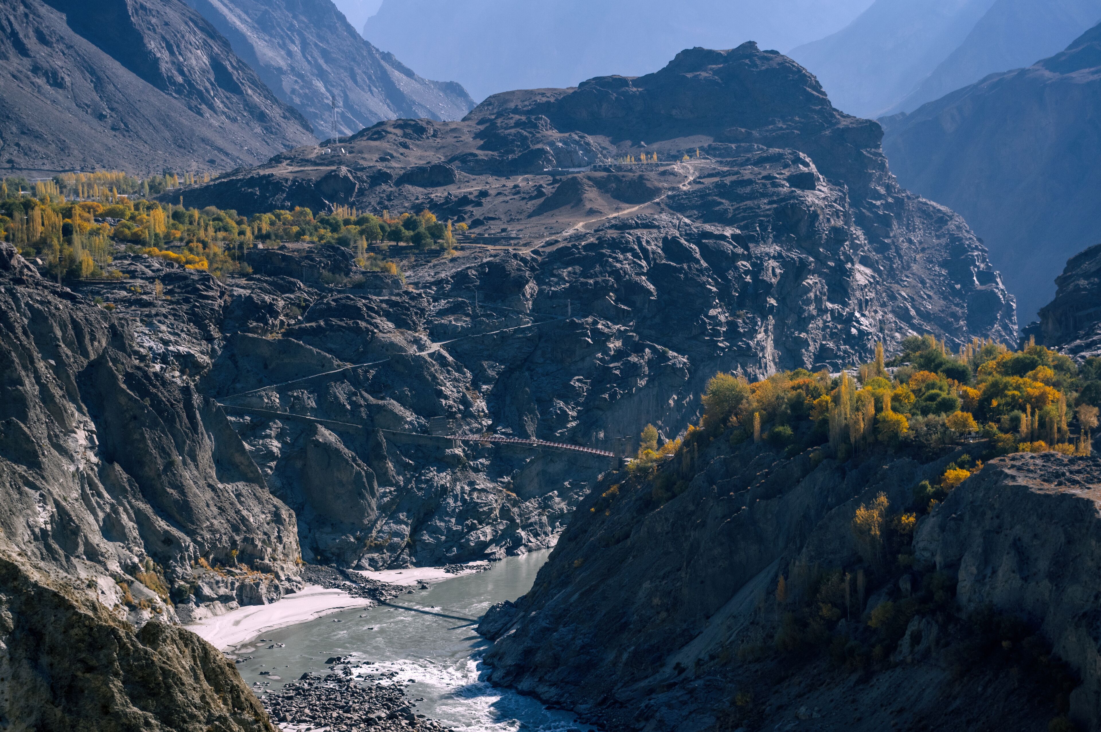 View of rugged mountains loom over a river valley, with a small bridge connecting the steep slopes under a clear sky, Jaglot, Gilgit Baltistan, Pakistan.