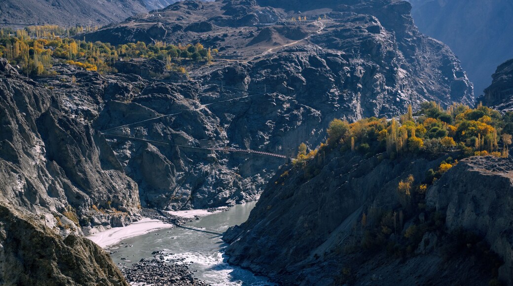 View of rugged mountains loom over a river valley, with a small bridge connecting the steep slopes under a clear sky, Jaglot, Gilgit Baltistan, Pakistan.