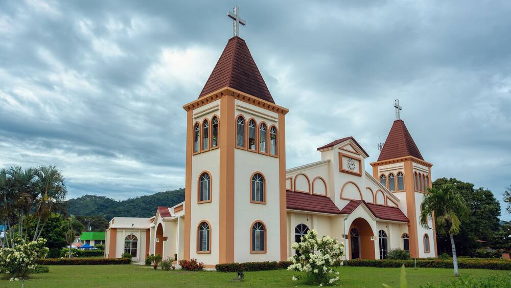 A church with two towers and a cross on top, surrounded by greenery and palm trees.