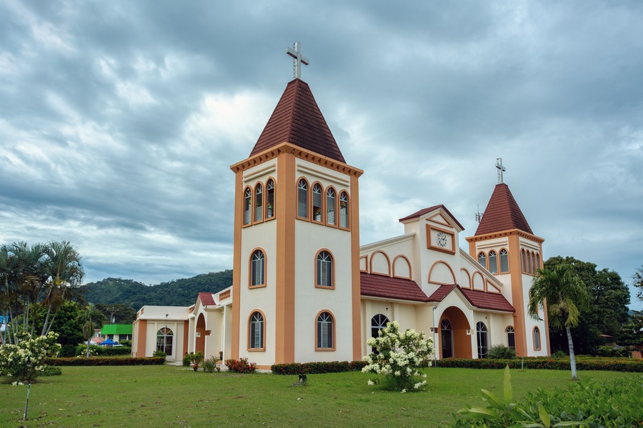 Una iglesia con dos torres y una cruz en la parte superior, rodeada de vegetación y palmeras.