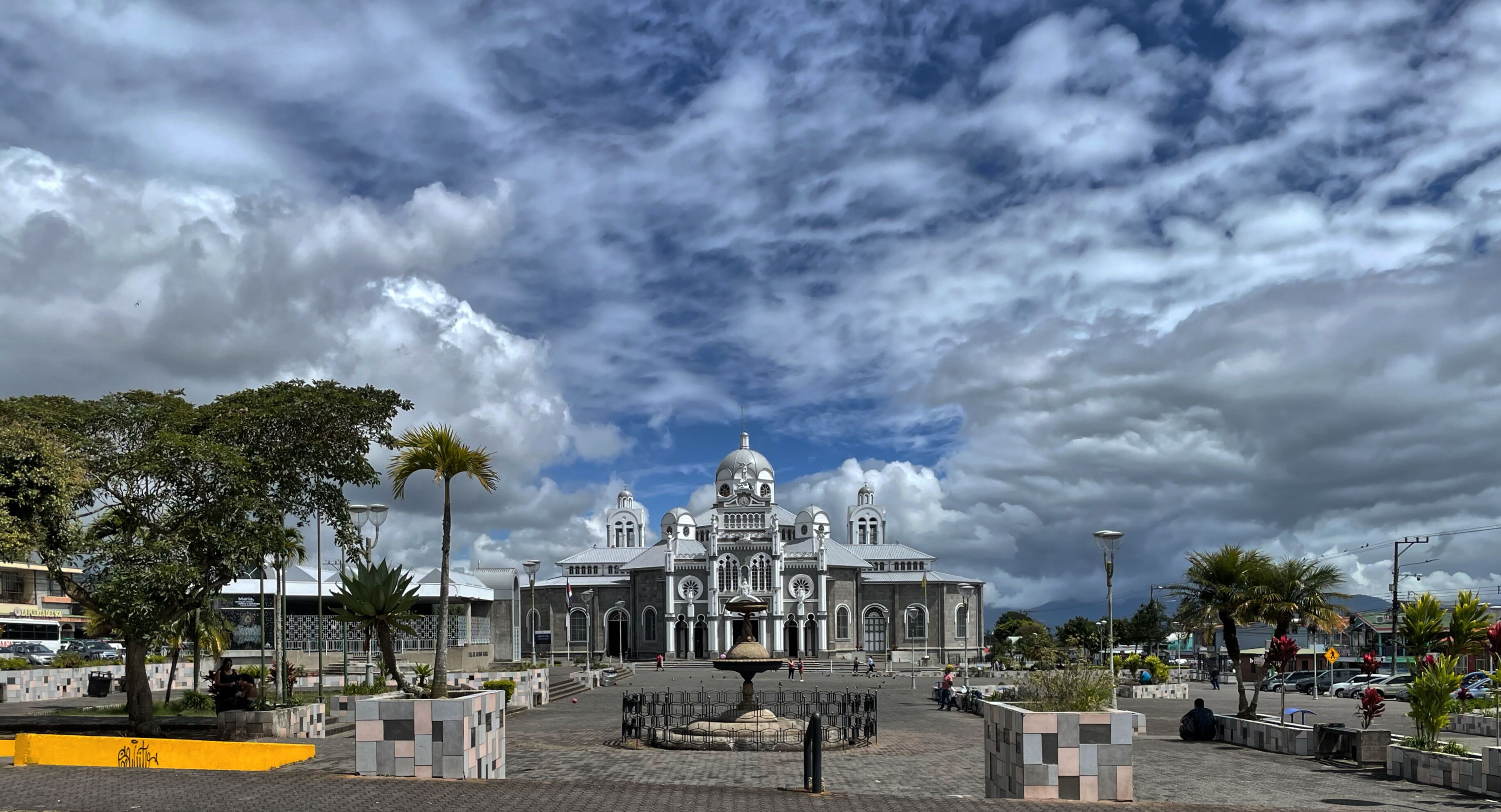 A grand building with domes and arches, surrounded by a plaza with a fountain and palm trees.