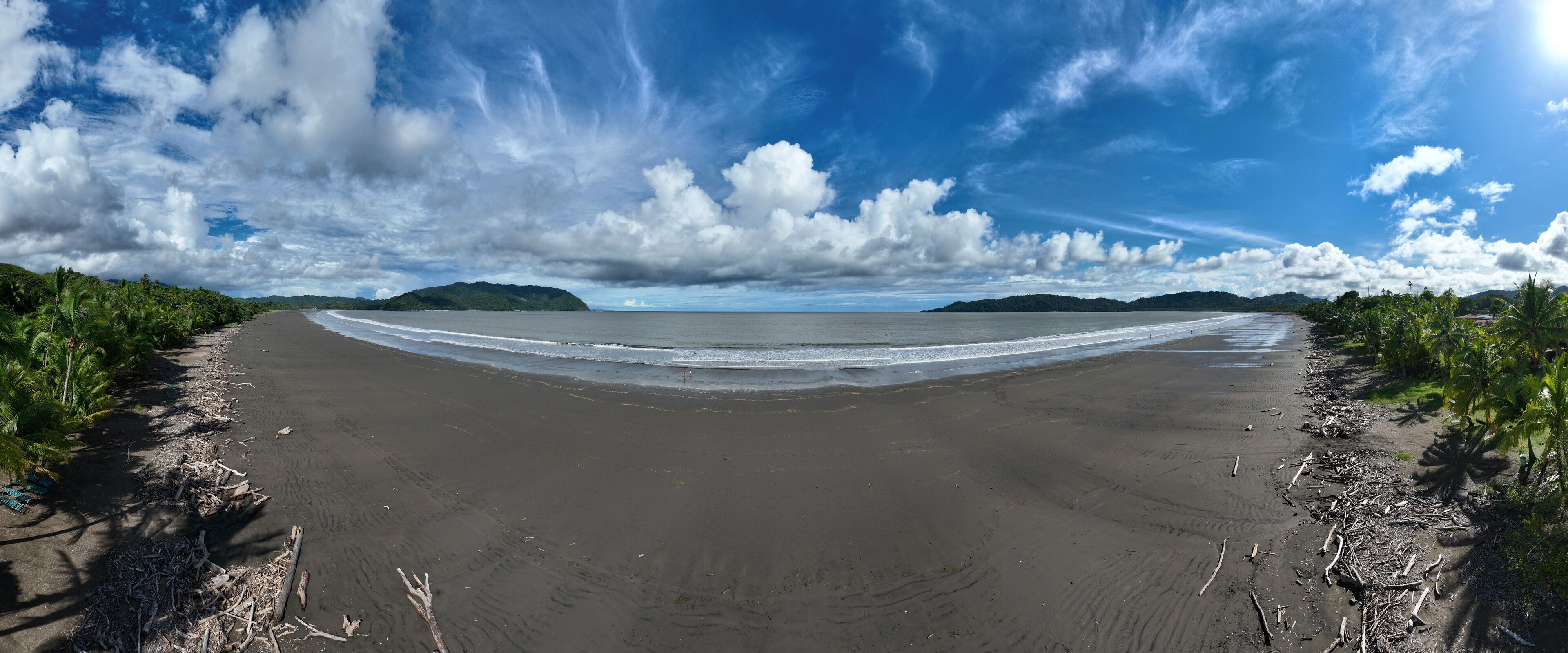 A beach with dark sand, palm trees, and a distant mountain range under a partly cloudy sky.