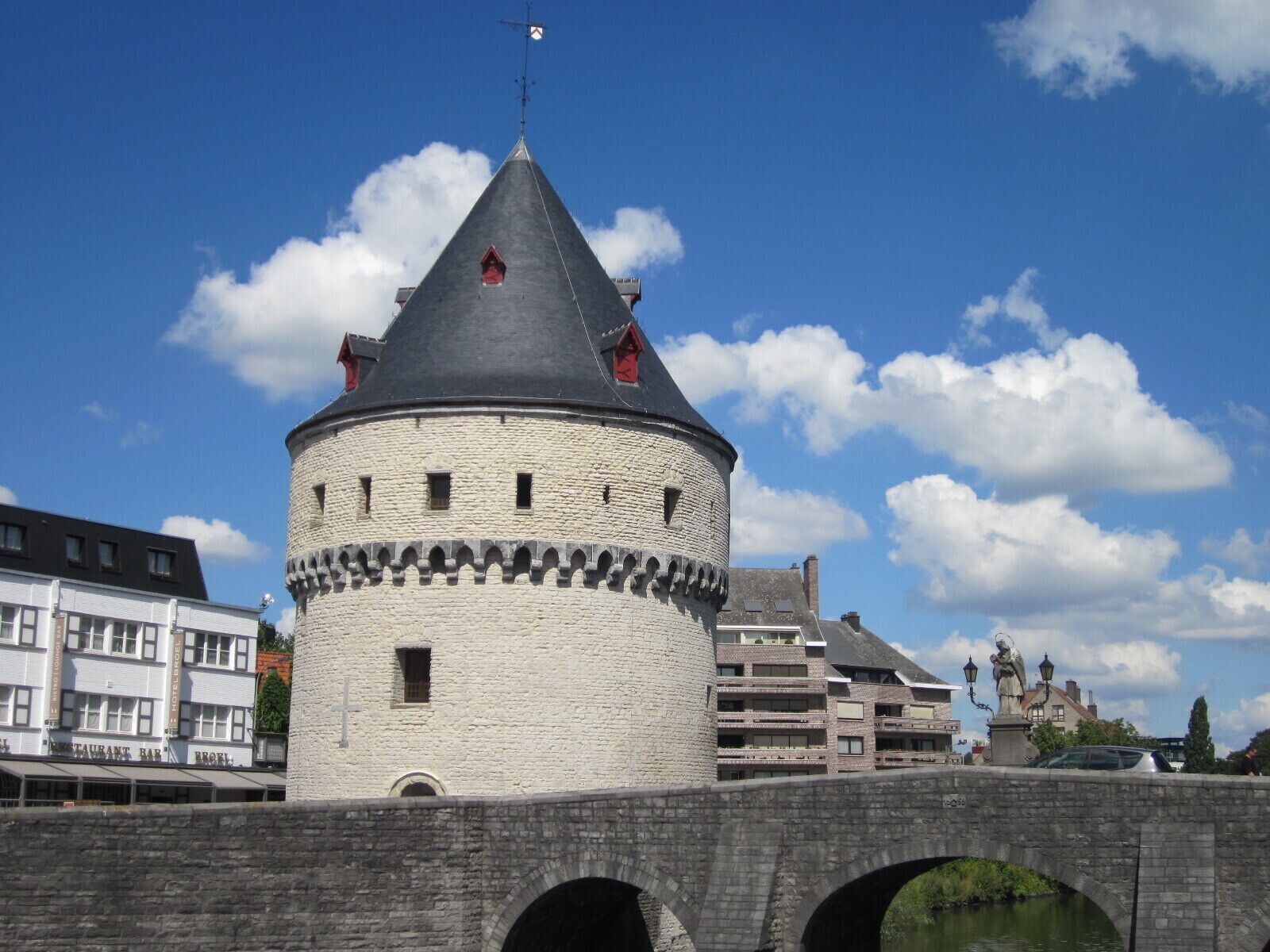 A historic stone tower with a conical roof, a bridge with arches, and modern buildings in the background.