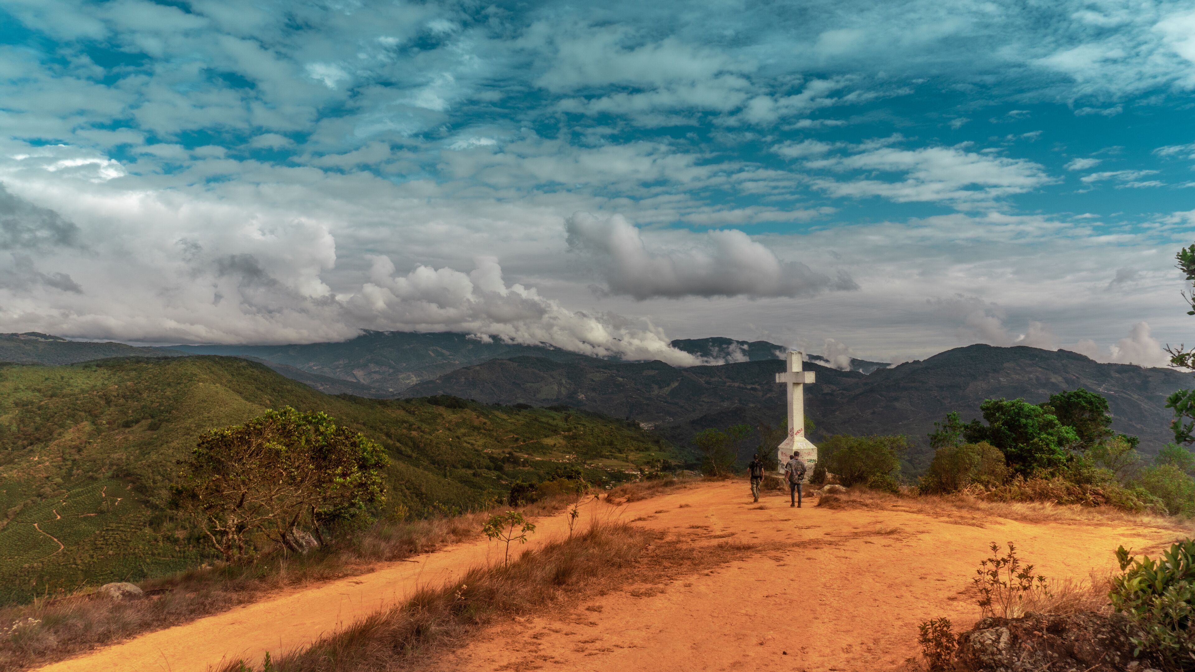 A cross on a hilltop with a dirt path leading up to it, surrounded by green hills and a cloudy sky.