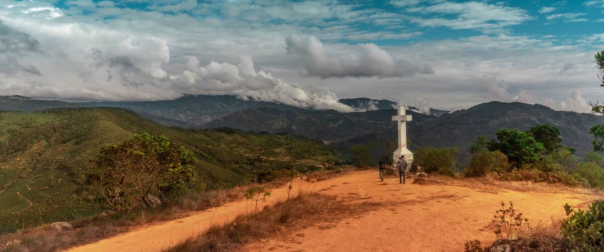 A cross on a hilltop with a dirt path leading up to it, surrounded by green hills and a cloudy sky.