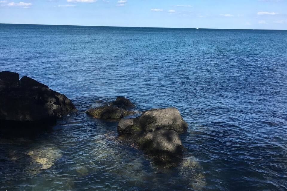 Mar azul cristalino com rochas visíveis e céu parcialmente nublado.