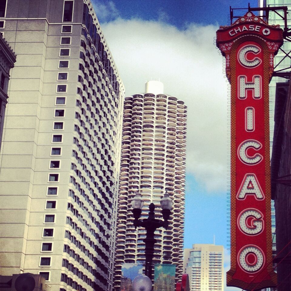 Paisagem urbana com um letreiro vermelho em destaque com a palavra 'CHICAGO' e um edifício alto com um design cilíndrico singular.