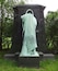 A statue of a figure draped in a cloak, standing on a pedestal in a grassy area with trees in the background.