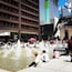 A public square with fountains, people sitting and walking, and modern buildings in the background.