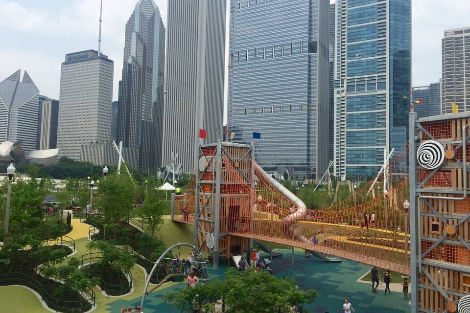 Une aire de jeux dotée de structures d’escalade et de toboggans, entourée d’un parc arboré, avec une vue sur la ville en arrière-plan.