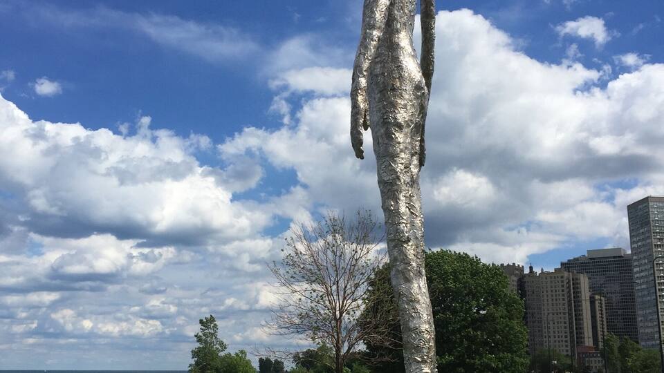 A tall metallic sculpture in a park with a clear sky and distant buildings.