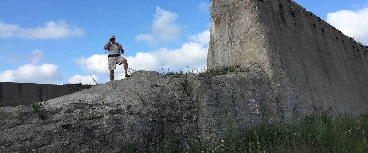 A man standing on a rocky outcrop with a large concrete structure in the background.