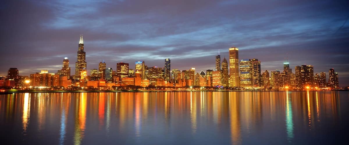 A city skyline at dusk with illuminated buildings reflecting on the water.