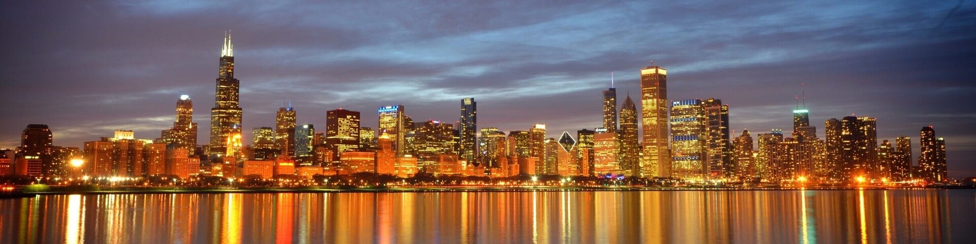 A city skyline at dusk with illuminated buildings reflecting on the water.