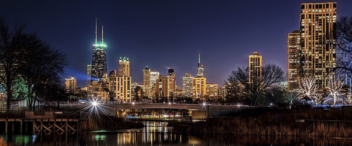 A nighttime city skyline reflected in a calm body of water, with illuminated buildings and a bridge.