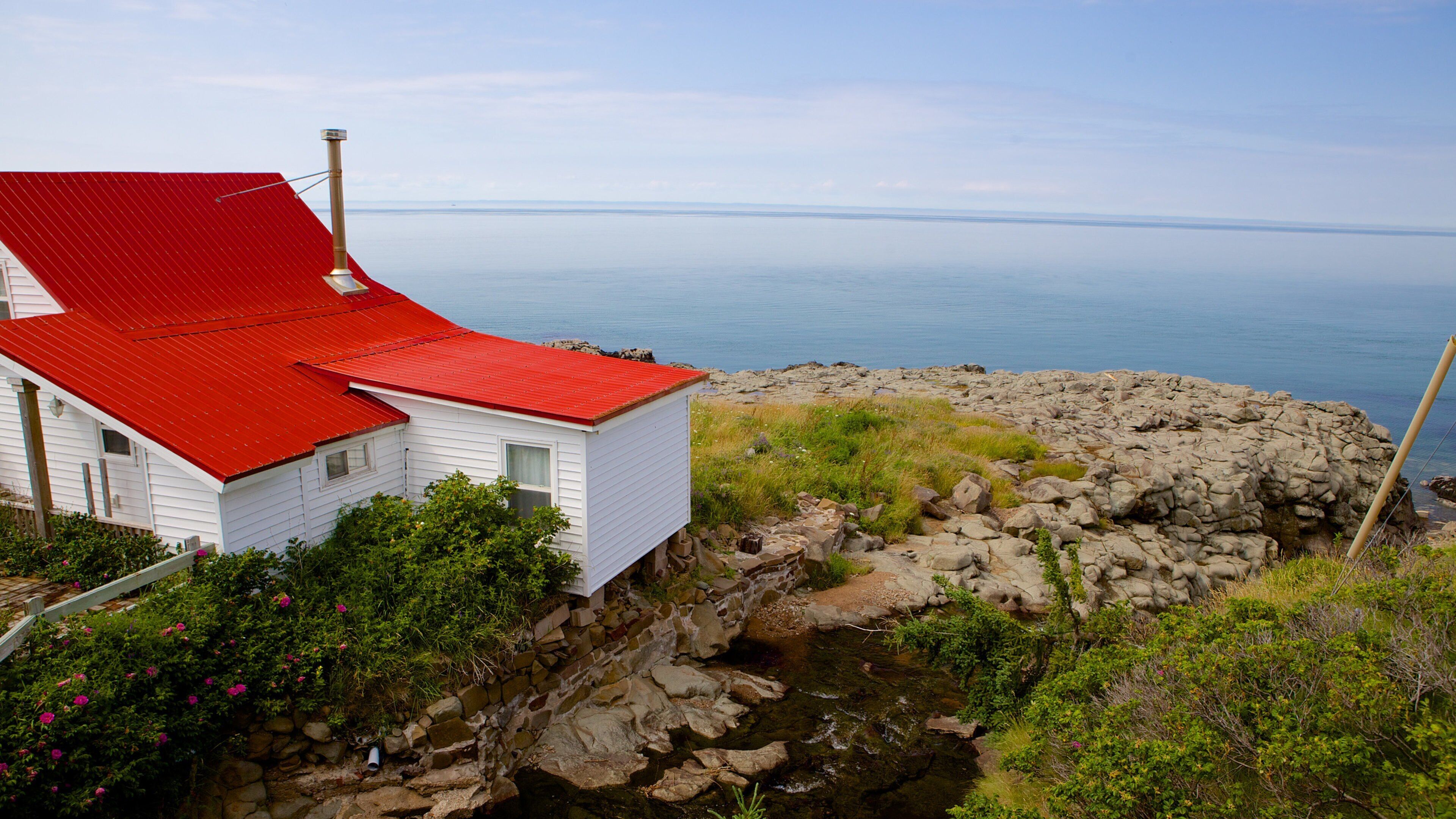 A white house with a red roof by a rocky shoreline with a view of the ocean.