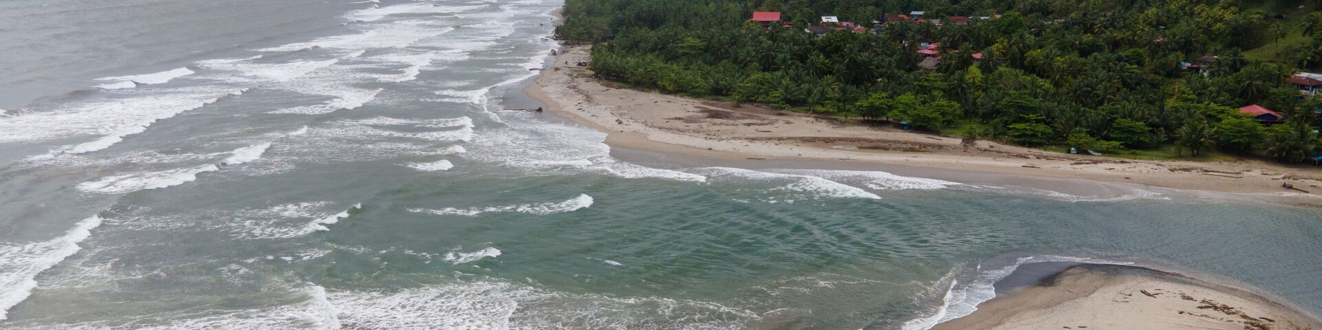 Paisaje costero con olas rompiendo sobre una playa arenosa, una ladera boscosa y una cadena montañosa distante bajo un cielo nublado.
