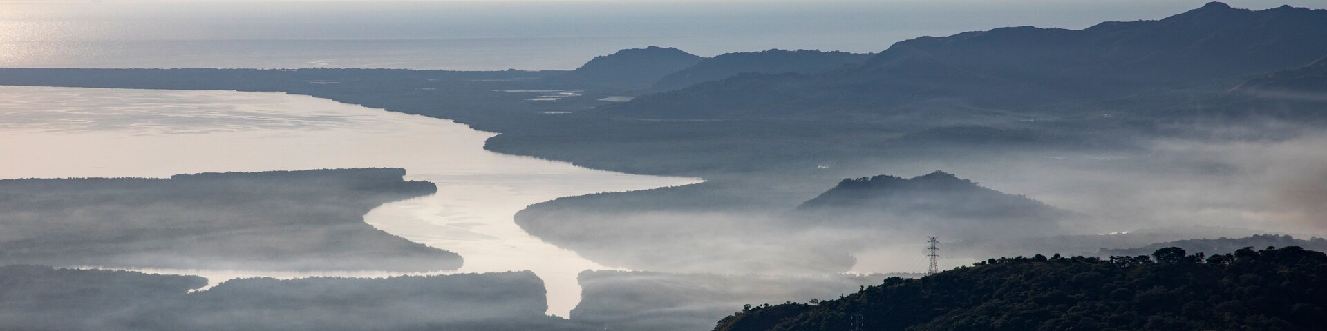 Paisagem costeira com um rio serpenteando por um vale, rodeado por colinas e névoa.