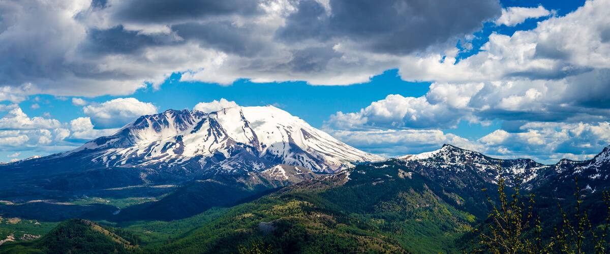 A snow-capped mountain range with a prominent peak, lush green valleys, and a partly cloudy sky.