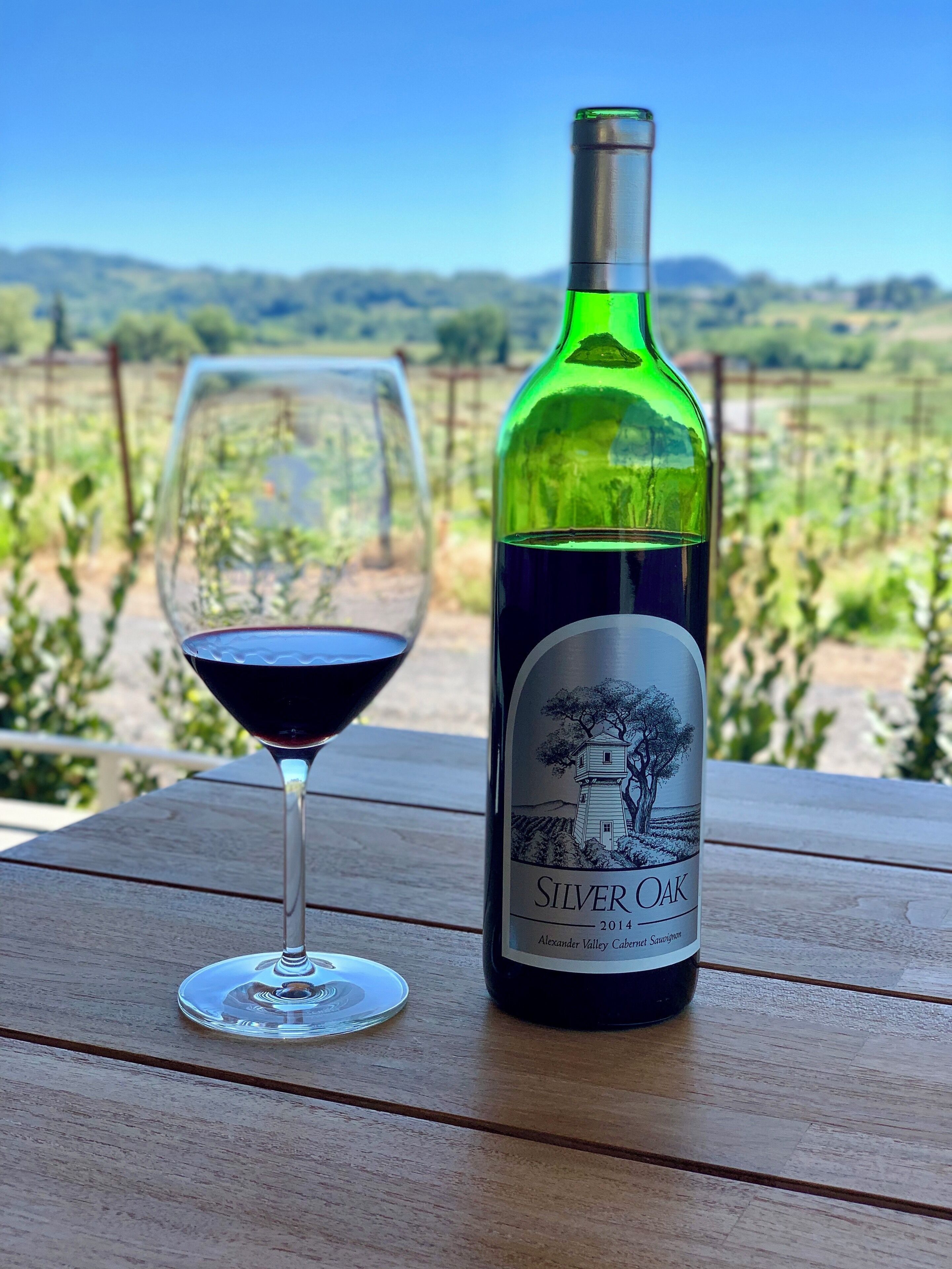 A bottle of Silver Oak wine and a glass of red wine on a wooden table with a vineyard in the background.