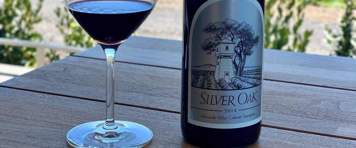 Una ampolla de vi Silver Oak i un got de vi vermell damunt d'un taula de fusta, amb un viñedo al fons.
