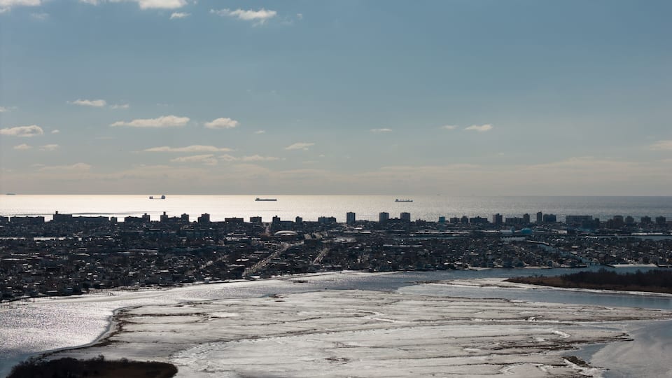A coastal cityscape with a frozen river in the foreground and ships on the distant water.