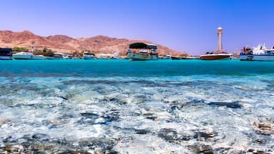 A clear turquoise sea with boats, a mountain range, and a tower in the background.