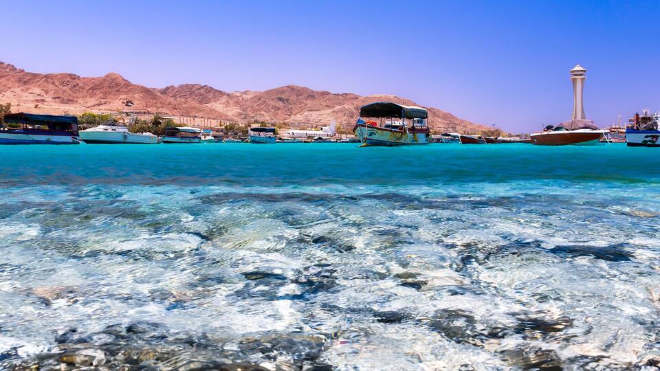A clear turquoise sea with boats, a mountain range, and a tower in the background.