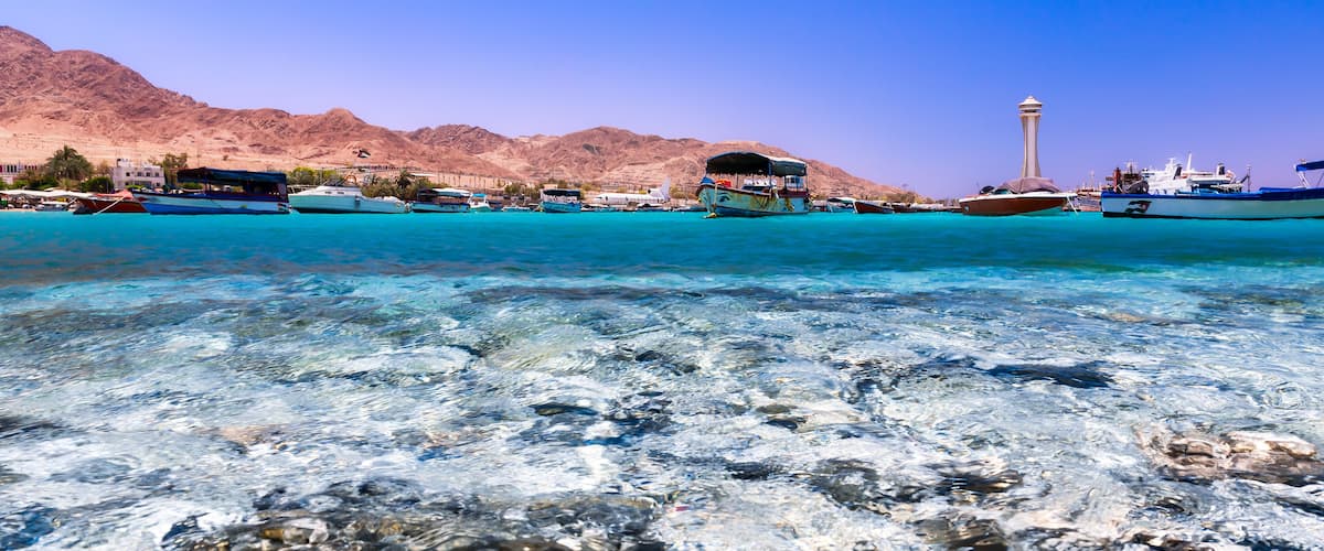 A clear turquoise sea with boats, a mountain range, and a tower in the background.