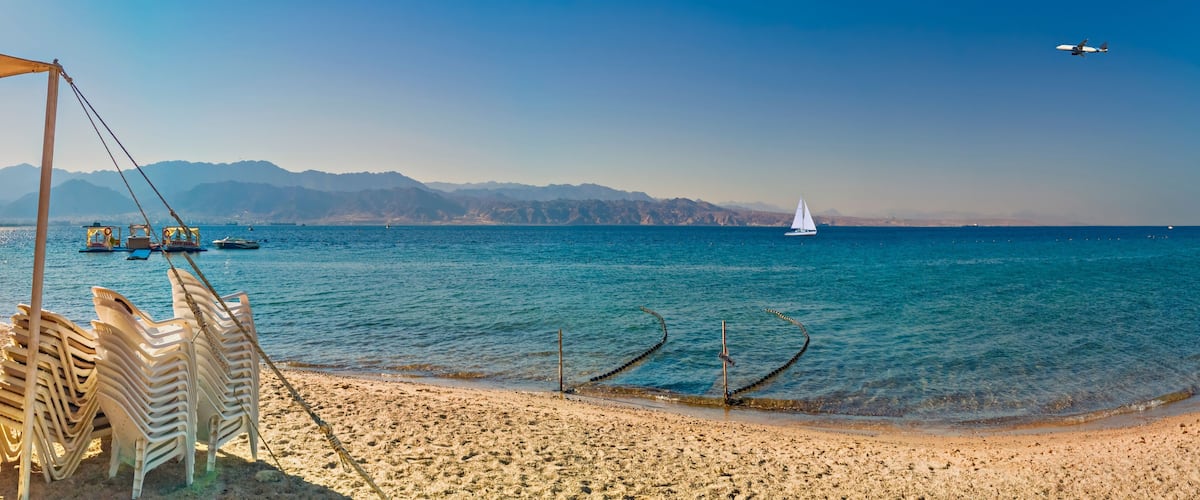 A beach with clear blue water, a sailboat, and mountains in the background.