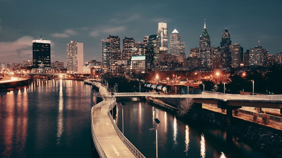 A nighttime cityscape with a river reflecting lights, a bridge, and various illuminated buildings.