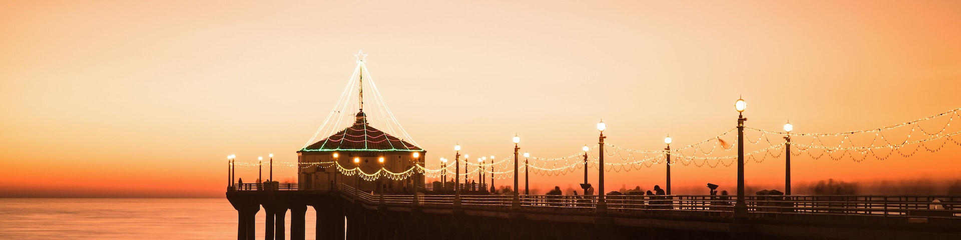 Une jetée surmontée d’une structure en forme de dôme, illuminée de guirlandes lumineuses, avec un coucher de soleil se reflétant sur l’eau.