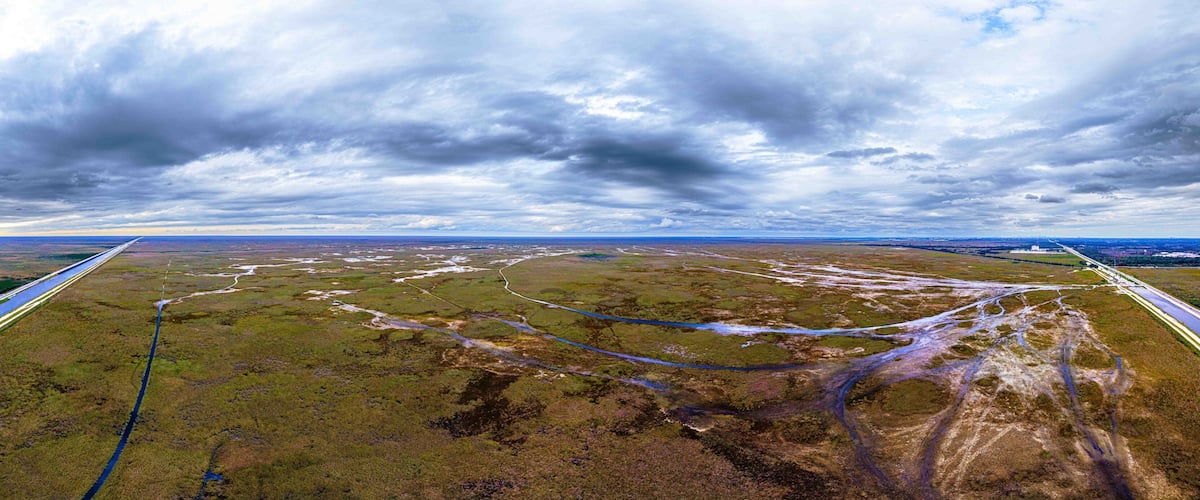 A vast landscape with a river, a road, and a distant city skyline under a cloudy sky.