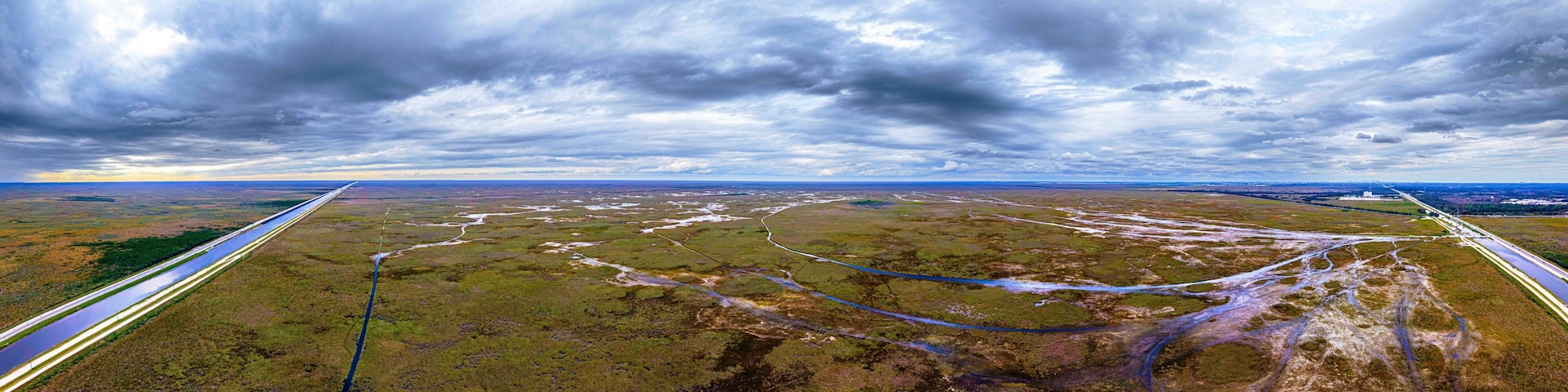 A vast landscape with a river, a road, and a distant city skyline under a cloudy sky.