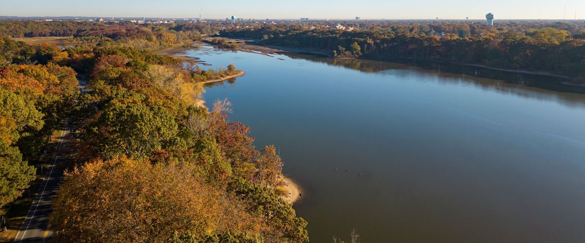 A river winding through a forested area with autumn foliage, a road parallel to the river, and distant cityscape.