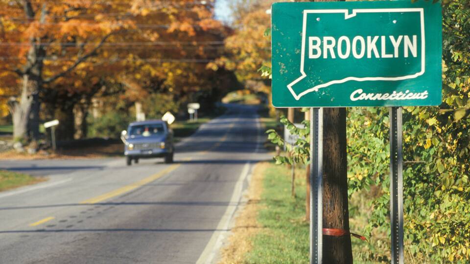 A road sign for Brooklyn, Connecticut, with autumn trees in the background.