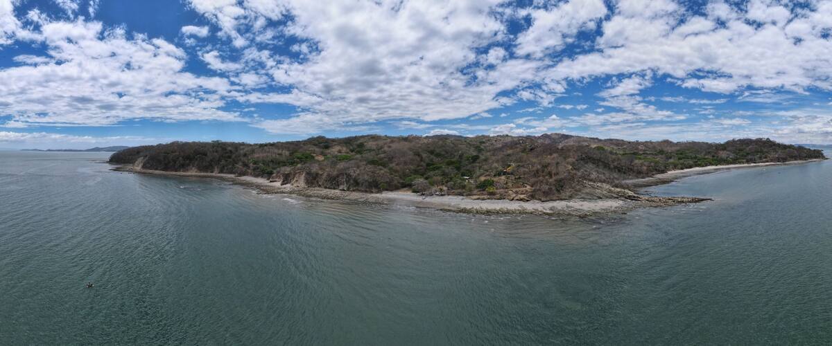 An aerial view of a small island with dense vegetation and a clear blue sky with scattered clouds.
