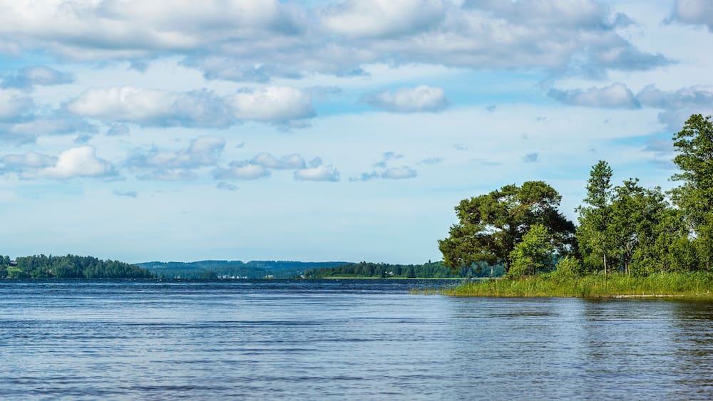 Ein ruhiger See mit bewaldeter Uferlinie und sanften Hügeln im Hintergrund, unter einem teils bewölkten Himmel.